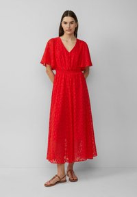 Vrouw staat tegen een grijze muur en draagt een rode V-hals midi-jurk met ﬂaddermouwen en bruine platte sandalen, met haar handen achter haar rug.