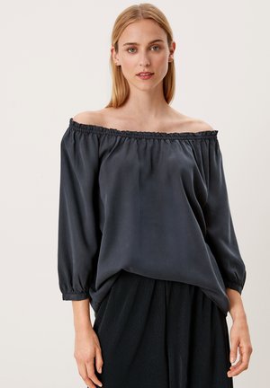 Tânără îmbrăcată într-o bluză întunecată, cu umărul descoperit, cu mâneci până la trei sferturi și o fustă plisată neagră, stând în fața unui fundal simplu.