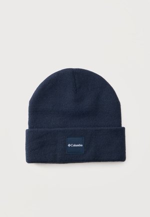 Gorro tejido de color azul oscuro con borde doblado y una etiqueta de la marca Columbia centrada en el puño sobre un fondo liso y claro.