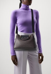 Sac à main à motifs géométriques marron avec un rabat en cuir noir et des finitions en or, porté en bandoulière sur un col roulé violet et un pantalon blanc.