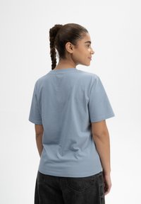 Hellesblaues, kurzärmliges T-Shirt aus Baumwolle. Glatte Textur, lockere Passform, runder Ausschnitt, von hinten gezeigt. Schwarze Hose sichtbar.