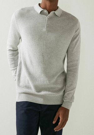 Mann trägt ein hellgraues, strukturiertes Langarm-Poloshirt und dunkle Hose, steht mit einer Hand hinter dem Rücken vor einfachem Hintergrund.