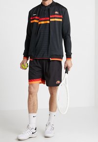 Man i svart tennisoutfit med röda och orangea ränder som håller i en tennisboll och en vit racket mot en enfärgad bakgrund.