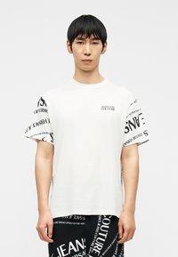 Ασιάτης άνδρας με μαύρα μαλλιά, ντυμένος με λευκό T-shirt Versace Jeans Couture και μαύρο σορτς με μεγάλα λευκά γράμματα, στέκεται μπροστά σε λευκό φόντο.