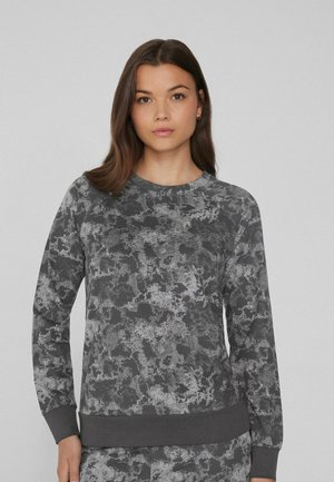 Jeune femme aux longs cheveux bruns portant un sweat-shirt à manches longues gris foncé et gris clair à motifs, sur un fond uni.