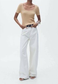 Mujer que lleva una camiseta de manga corta beige con canalé, pantalones blancos de pierna ancha de cintura alta, cinturón negro, pulseras plateadas y sandalias blancas.