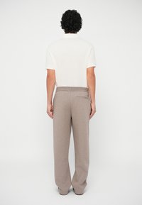 Vīrietis ar cirtainiem matiem stāv vērsts prom, tērpies baltā īsajās piedurknēs polo kreklā un brīviem beigu toņa biksēm uz neitrāla fona.