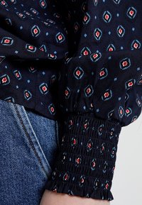 Blusa blu navy con motivo a diamante e floreale in rosso e blu. Presenta un polsino arricciato con bordi sfrangiati e tessuto leggero.