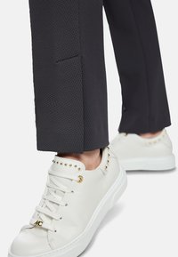 Weiße Ledersneaker mit goldenen Nieten an den Seiten, kombiniert mit strukturierten dunkelgrauen Hosen mit seitlichem Schlitz.