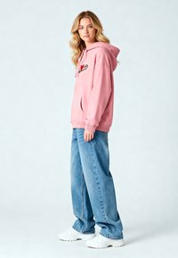 Felpa rosa con una grande tasca frontale, design con patch ricamato. Abbinata a jeans larghi di un blu chiaro e sneakers bianche.