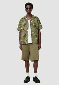 Jovem usando óculos de sol, camisa de manga curta com estampa de palmeiras verdes por cima de uma t-shirt branca, calções verde-oliva, meias brancas e sapatos pretos, em pé.