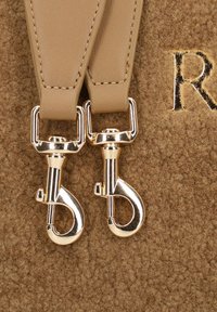 Twee beige leren tasriemen met gouden metalen draaiklinken op een textuur van bruin stof met gedeeltelijk gestikte letters.