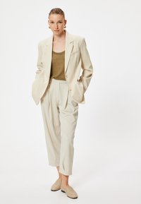 Completo beige su misura con blazer a un bottone e pantaloni plissettati, abbinato a una canotta oliva. Tessuto morbido e linee pulite.