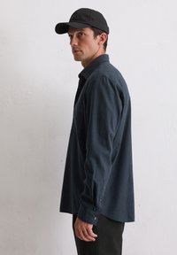 Donkerblauw geruit overhemd met een zak, lange mouwen en knoopsmanchetten, gecombineerd met een zwarte pet en donkere broek.
