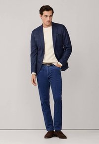 Tmavě modrý kostkovaný blejzr přes krémový svetr, spárovaný s modrými kalhotami a hnědými botami. Blejzr má klopový límec s výbrusem a kapesníček.