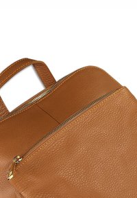 Bruine, gestructureerde leren tas met een gouden rits die gedeeltelijk openstaat, waardoor de binnenkant en de korte handgreep zichtbaar zijn tegen een witte achtergrond.