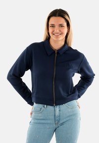 Giacca a zip blu navy con colletto strutturato, orlo a coste e logo in rilievo sul lato sinistro del petto, abbinata a jeans blu chiaro.