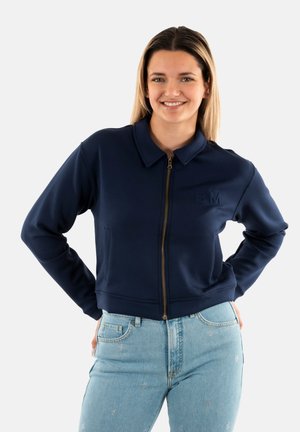 Veste zippée bleu marine avec un col structuré, un ourlet côtelé et un logo embossé sur la poitrine gauche, assortie avec un jean bleu clair.