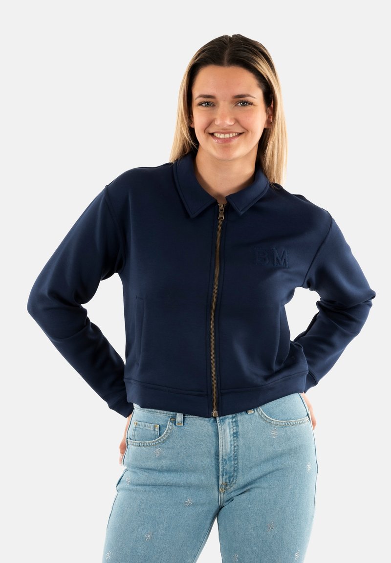 Giacca a zip blu navy con colletto strutturato, orlo a coste e logo in rilievo sul lato sinistro del petto, abbinata a jeans blu chiaro.