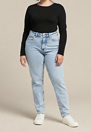 Vrouw draagt lichtblauwe jeans met hoge taille, zwart shirt met lange mouwen en witte instapschoenen, staand tegen een effen beige achtergrond.