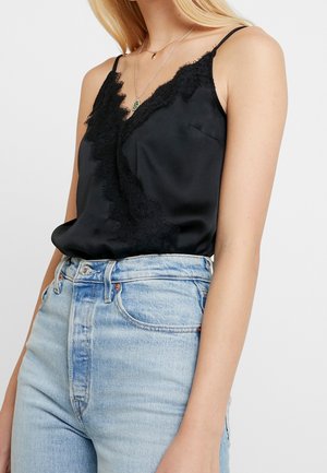 Mujer con un camisón de satén negro con encaje, metido dentro de unos vaqueros azul claro de talle alto, con dos collares dorados en capas.