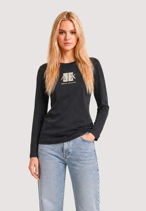 Blondynka ubrana w czarną koszulkę z długim rękawem marki Armani Exchange i jasnoniebieskie jeansy, stoi z jedną ręką w kieszeni.