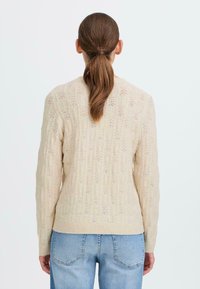 Beige strikkegenser med teksturert mønster, med rund halslinning og lange, tettsittende ermer. Synlig ribbestrikket kant ved midjen.