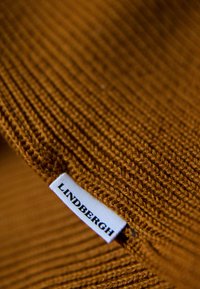 Tissu tricoté côtelé marron avec une étiquette blanche portant le texte "LINDBERGH" en noir. La texture est douce avec des détails de couture visibles.