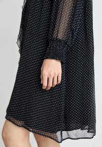 Schwarzes Polka-Dot-Kleid mit durchsichtigen langen Ärmeln, gesammelten Manschetten und fließendem Saum. Der Stoff hat eine glatte Textur und wirkt leicht.