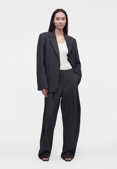 Femme debout dans une pose détendue, portant un costume à rayures ample, un débardeur blanc et des sandales sur un fond uni clair.