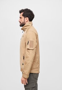 Beige zip-up jacket with a high collar, textured fabric, side pockets, and a patch pocket on the upper arm. Worn with gray cargo pants.
