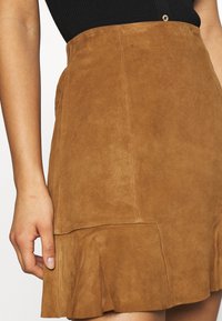 Jupe en daim marron avec un ourlet évasé, présentant une texture lisse et une taille ajustée. Détail d'un petit bouton doré en haut.