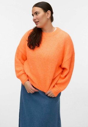 Ženska z dolgimi temnimi lasmi, oblečena v svetlo oranžen prevelik pulover in modro krilo, gleda levo proti enostavni ozadju.