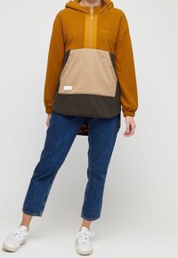 Kleurgeblokte hoodie in mosterd, beige en donkerbruin fleece; heeft een voorzak en een halve ritssluiting. Draagt met blauwe jeans.