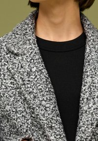 Abrigo texturizado en mezcla de lana gris y blanco con cuello de solapa, sobre una blusa negra de canalé, mostrando un diseño en capas.