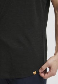 Tricou negru din material texturat, cu un patch mic în forma de pătrat la tiv, ținut de o mână aproape de cusătura laterală.