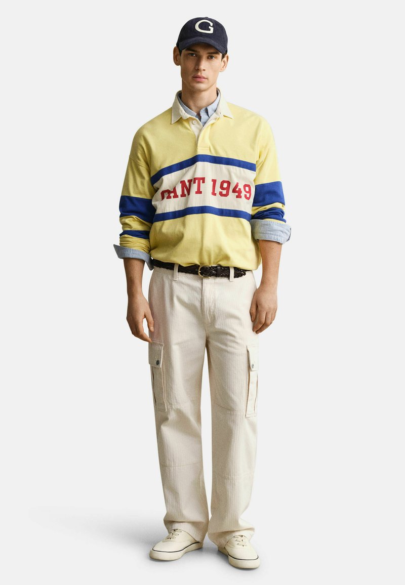 Jeune homme portant un maillot de rugby jaune avec l'inscription "GANT 1949", un pantalon cargo beige, des baskets blanches et une casquette bleu marine avec un "G" blanc.