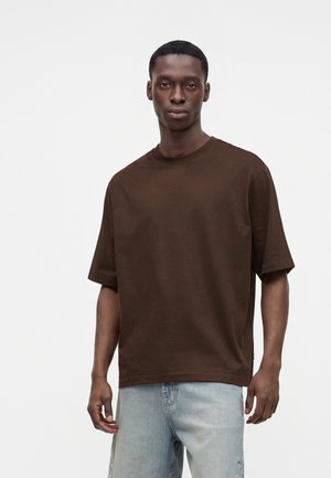 Jeune homme portant une chemise marron unie oversize à manches courtes et un jean bleu clair, debout devant un fond blanc.