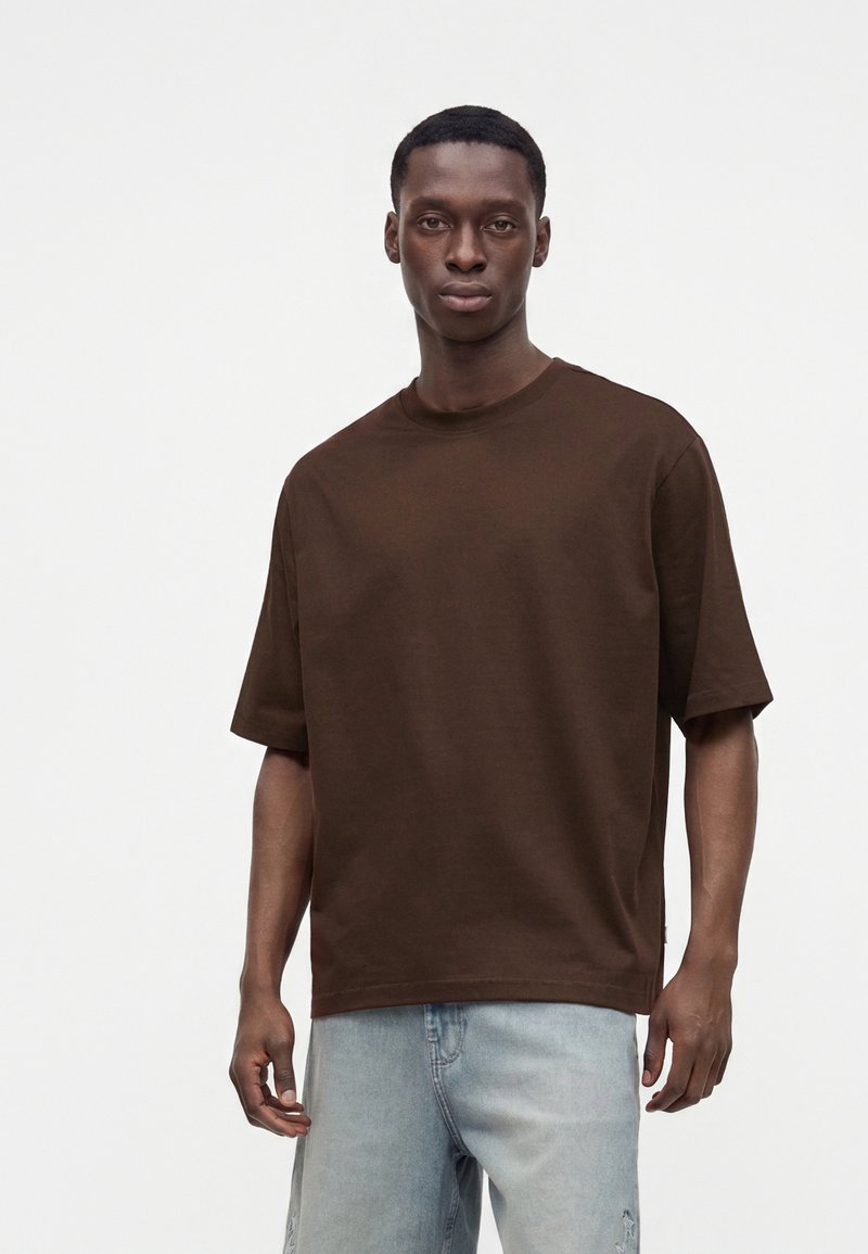Jeune homme portant une chemise marron unie oversize à manches courtes et un jean bleu clair, debout devant un fond blanc.
