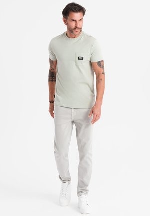 Camiseta de algodón verde claro con un bolsillo en el pecho, combinada con jeans gris claro y zapatillas blancas. Presenta mangas cortas y un escote redondo.