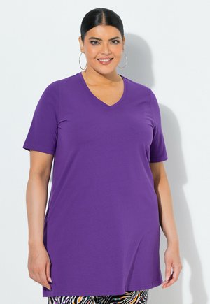 Uśmiechnięta kobieta o ciemnych włosach w kolczykach-kołach nosi fioletową tunikę z krótkim rękawem i dekoltem w serek oraz spodnie w wzory na białym tle.