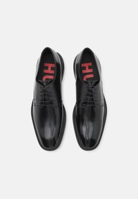 Chaussures habillées en cuir noir avec une finition brillante, à lacets, bout rond, et une semelle intérieure rembourrée arborant un logo rouge "HUH".
