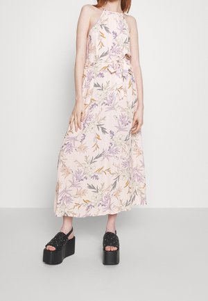Robe longue rose clair avec un motif floral en violet, orange et gris. Encolure dos-nu et détail ceinture nouée. Portée avec des sandales à plateforme noires.