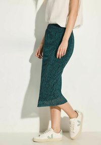Gonna midi teal con motivo geometrico, realizzata in tessuto leggero, abbinata a sneaker bianche con accenti verdi e un semplice design con lacci.