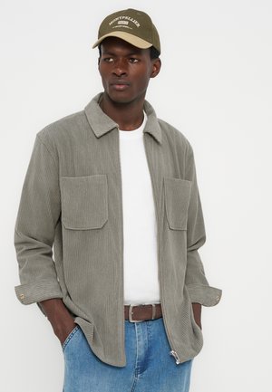 Homme portant une veste en velours côtelé vert olive sur une chemise blanche, un jean bleu et une casquette vert olive avec le logo de Montpellier, les mains dans les poches.