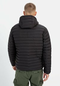 Sort puffet jakke med vandret quiltet design og hætte. Lavet af et letvægtsmateriale. Båret over grønne cargo bukser.