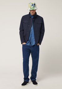 Marineblauwe jas met twee voorzakken, lichtblauw overhemden, blauwe jeans en een bloemenemmerhoed. Zwarte instap schoenen maken de outfit compleet.