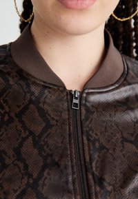 Rudas bombonešis su gyvatės odos raštu, turintis zip užsegimą ir raukšlėtą apykaklę. Lygus paviršius su blizgiais akcentais.