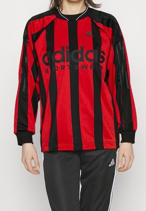 Person iført en rød og sort Adidas sportstrøje med lodrette striber, langærmet, og sorte Adidas bukser med hvide striber.