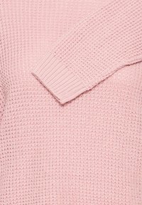 Manche de pull en tricot rose avec une bordure côtelée posée en diagonale sur un tissu corporel texturé assorti.